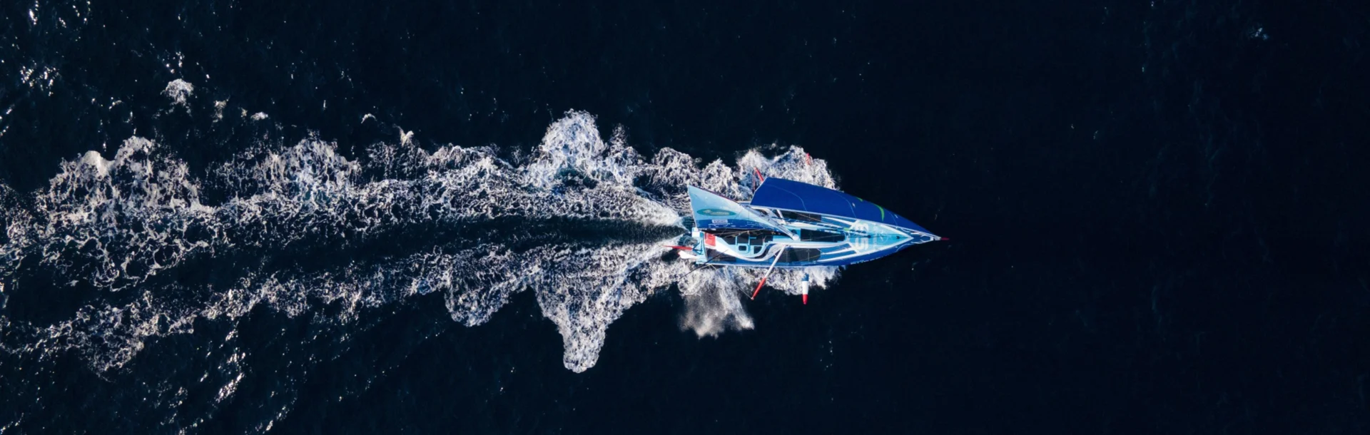 Vue du ciel de l'IMOCA Groupe Dubreuil 2, filant sur l'océan en laissant derrière lui un long sillage d'écume blanche.