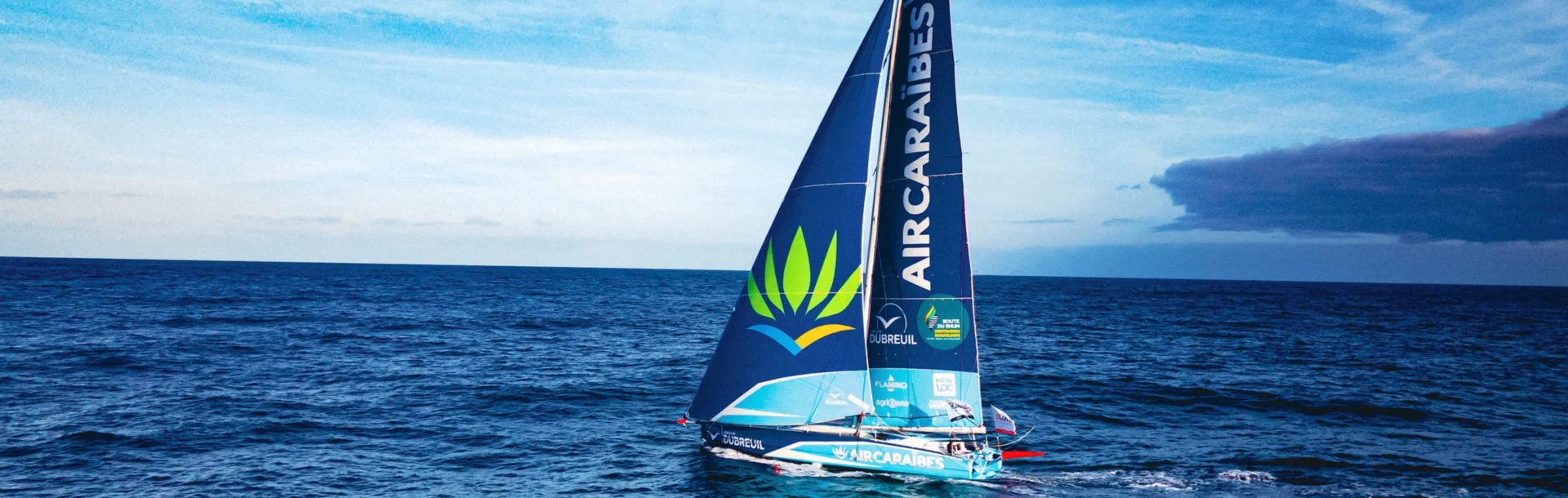 L'IMOCA Groupe Dubreuil 2 navigue au près sur l'océan, sa grand-voile et son foc arborant les logos Air Caraïbes et Groupe Dubreuil sous un ciel bleu.