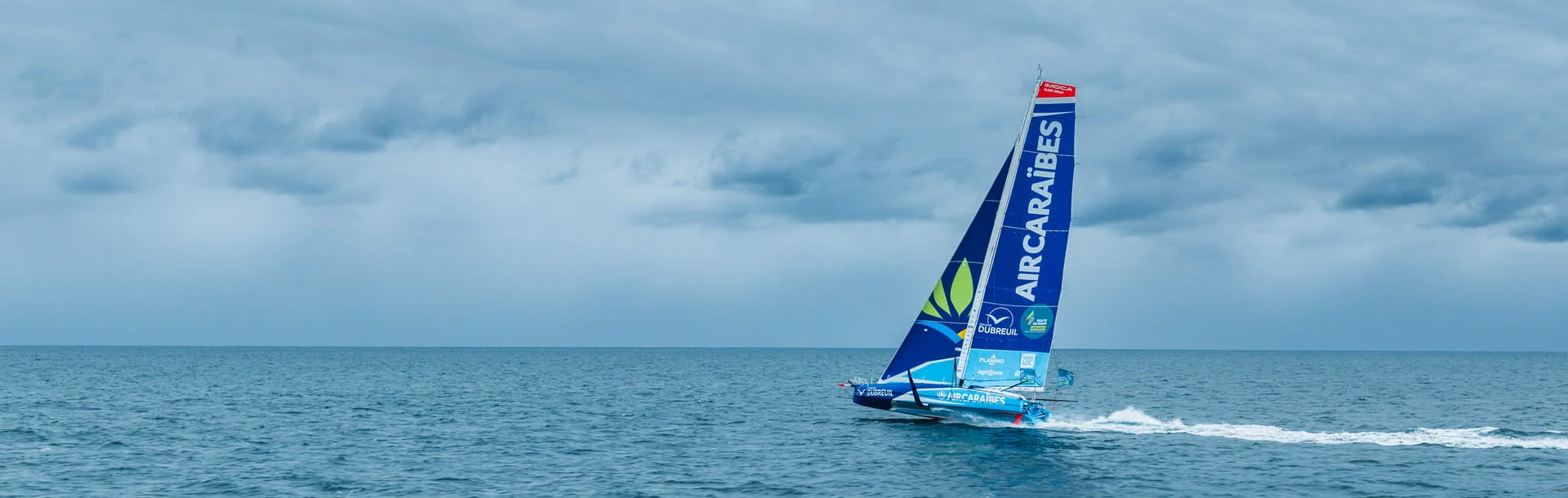 Voilier IMOCA Groupe Dubreuil – Air Caraïbes sur ses foils en mer, ciel nuageux